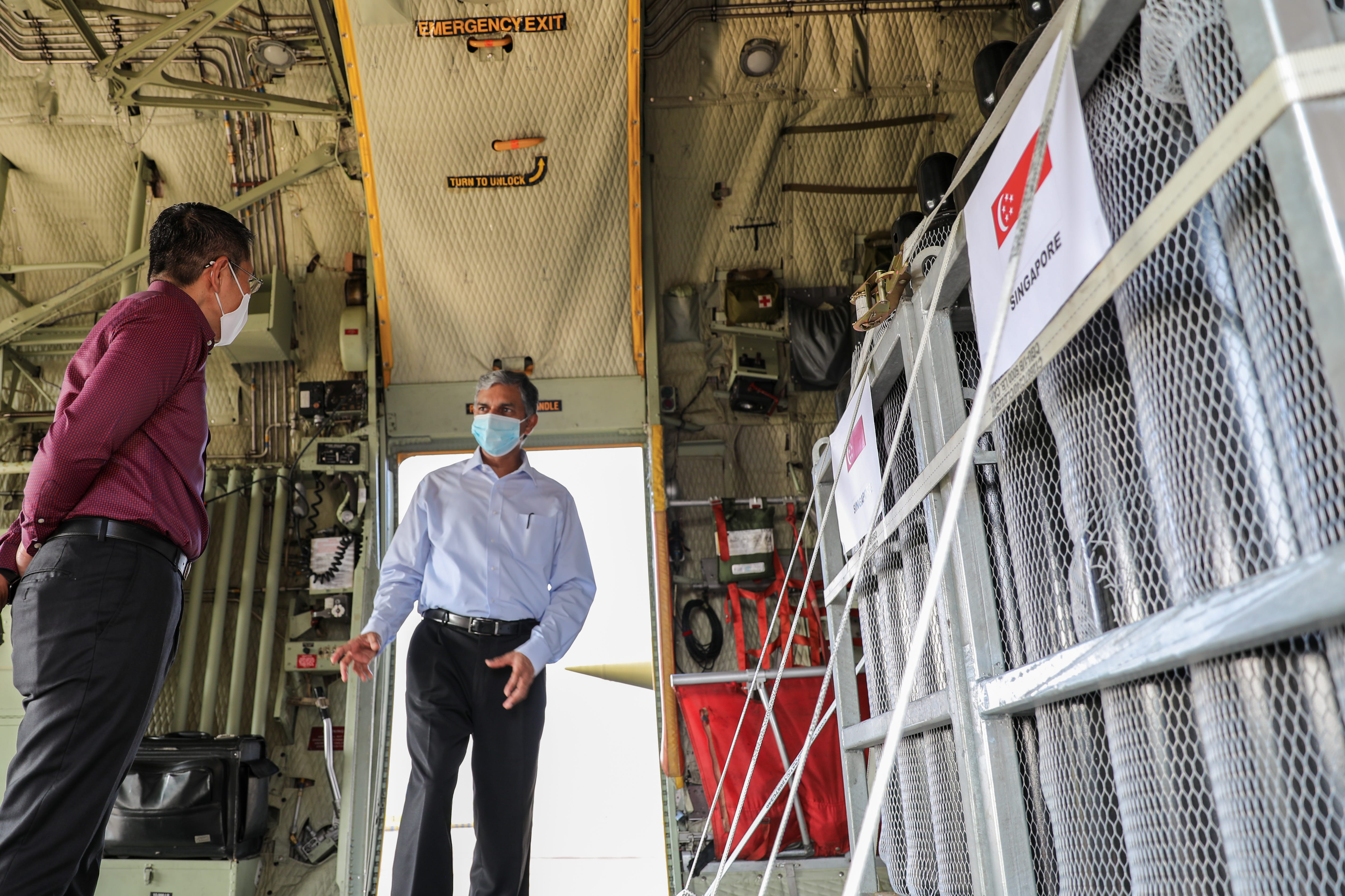 Inside military transport, two masked people conversing; cargo with Singapore flags visible.